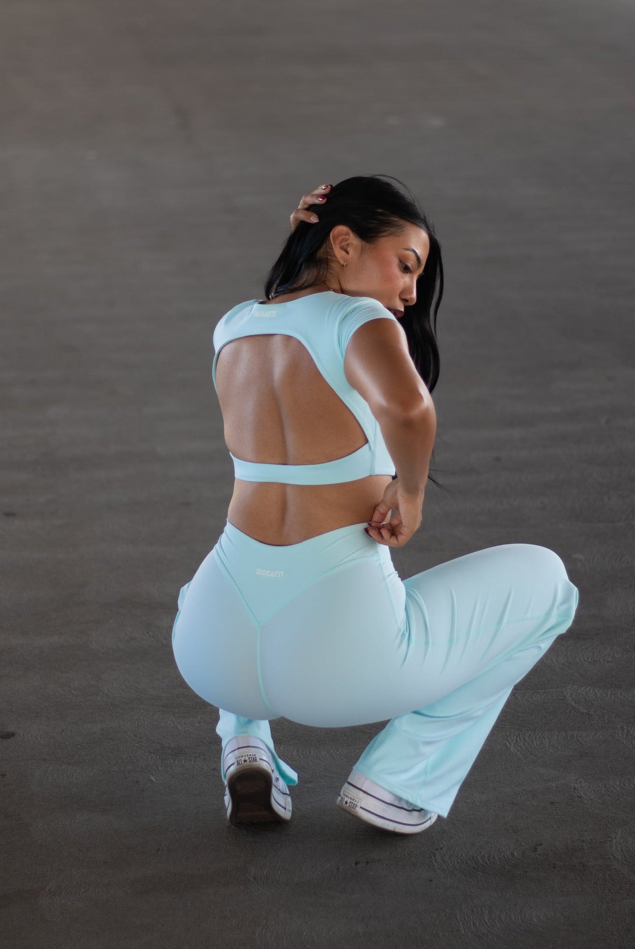 A woman in a light blue activewear set, featuring a strappy open-back sports bra and matching high-waisted pants, crouching in a spacious concrete parking structure.