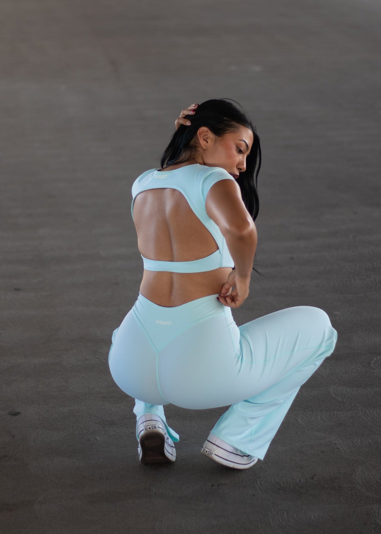 A woman in a light blue activewear set, featuring a strappy open-back sports bra and matching high-waisted pants, crouching in a spacious concrete parking structure.