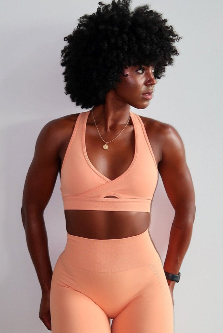 A woman wearing a blue sports bra with a deep V neck design and matching blue shorts. She is standing with her hands behind her head.