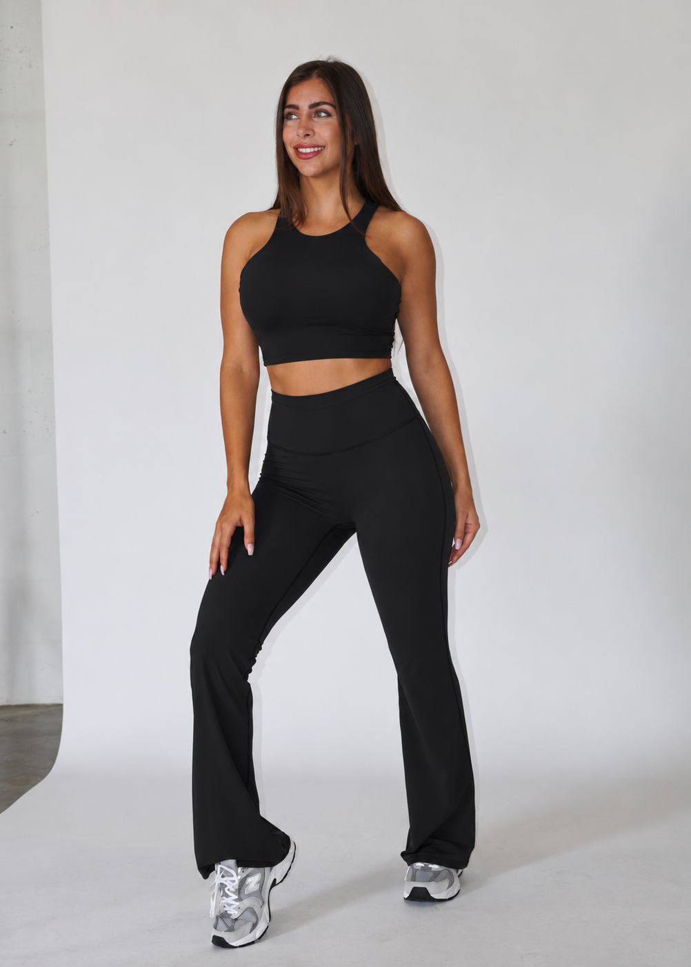 A woman modeling a black athletic two-piece set consisting of a halter crop top and high-waisted flared leggings, paired with silver sneakers, in a studio setting.