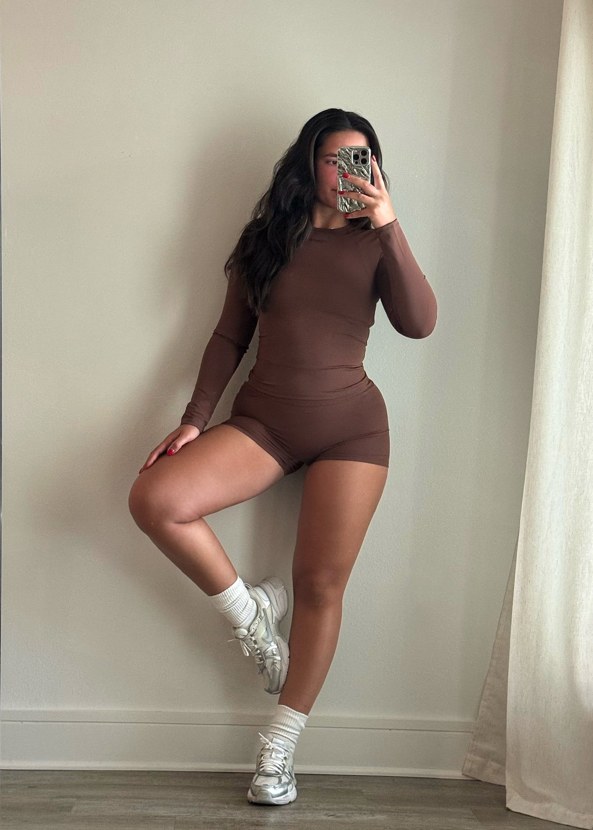 Person wearing a brown bodysuit taking a mirror selfie in a room with light-colored walls and curtains.