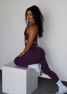 A woman with curly dark hair sits on a white cube in a studio setting, wearing a matching deep purple activewear set consisting of a sports bra and high-waisted leggings, paired with white socks and white sneakers.