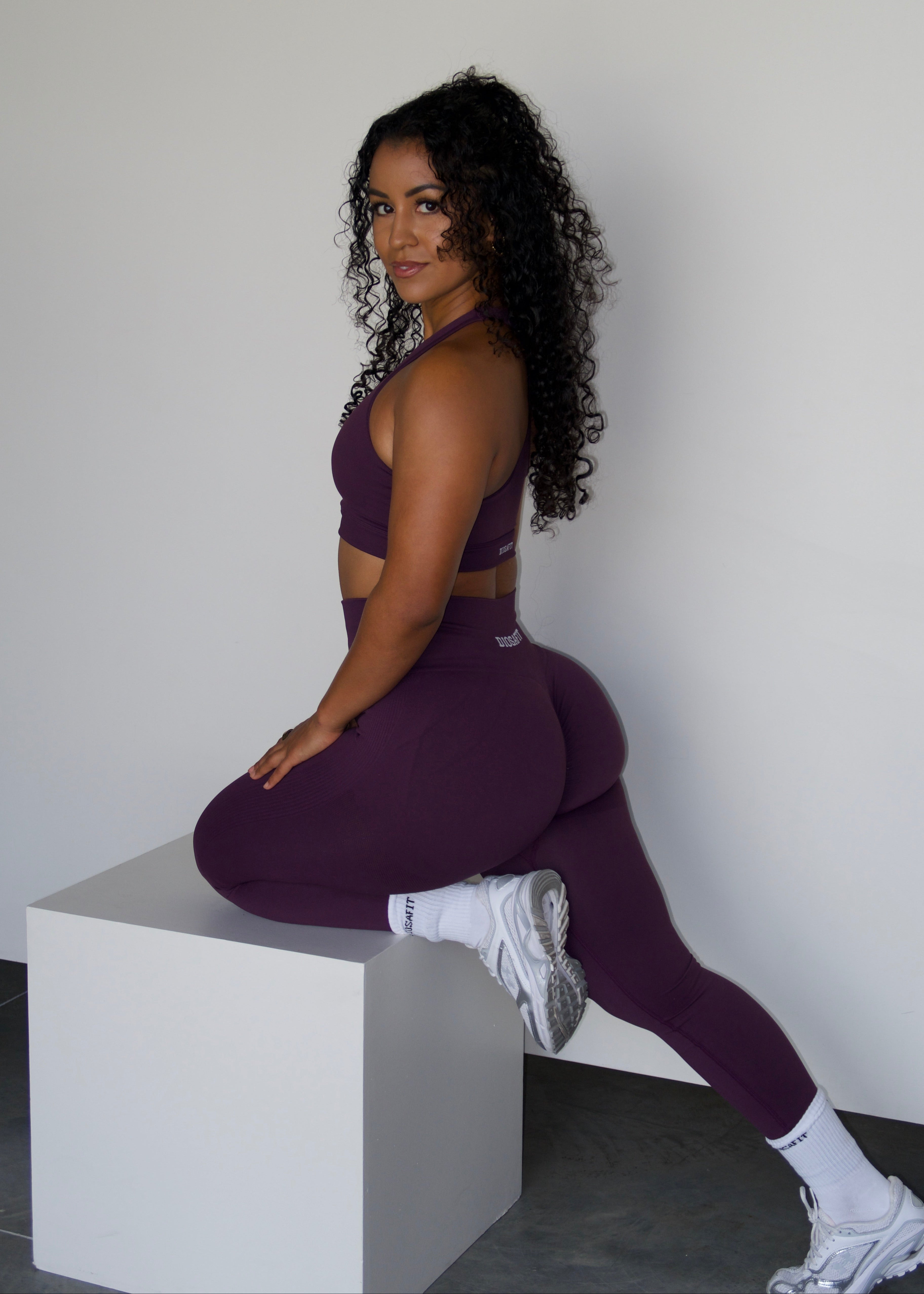 A woman with curly dark hair sits on a white cube in a studio setting, wearing a matching deep purple activewear set consisting of a sports bra and high-waisted leggings, paired with white socks and white sneakers.
