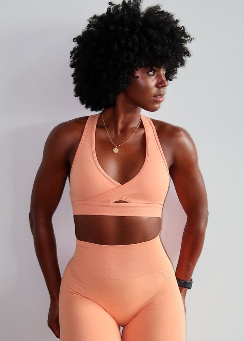 A woman wearing a blue sports bra with a deep V neck design and matching blue shorts. She is standing with her hands behind her head.