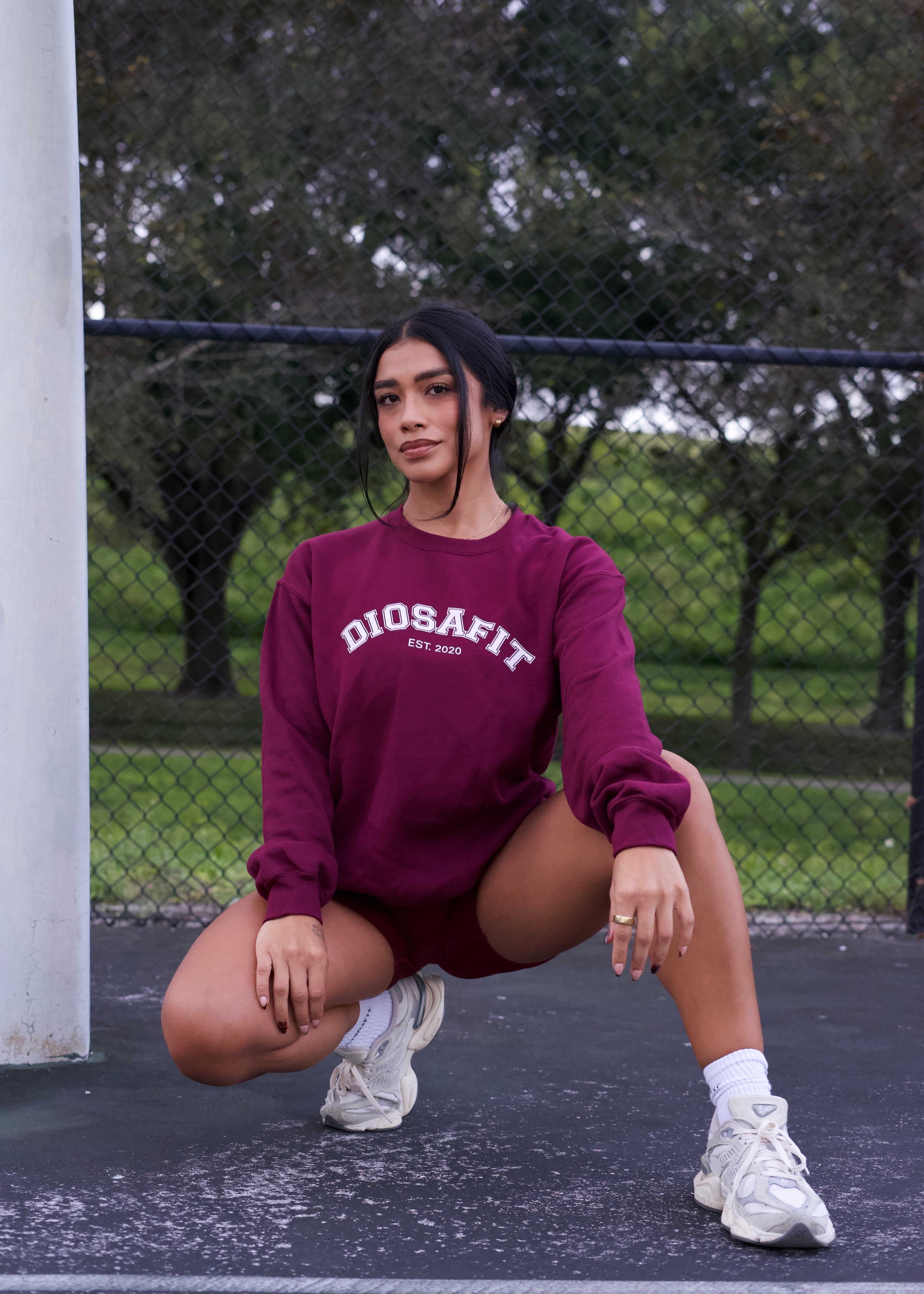 A woman crouching on an outdoor basketball court wearing a burgundy crewneck sweatshirt with 'DIOSAFIT EST. 2020' printed in white on the chest, paired with matching burgundy shorts and white sneakers.