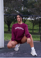 A woman crouching on an outdoor basketball court wearing a burgundy crewneck sweatshirt with 'DIOSAFIT EST. 2020' printed in white on the chest, paired with matching burgundy shorts and white sneakers.