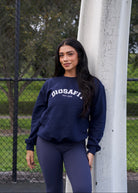 A woman stands outdoors leaning against a white pole, wearing a navy blue sweatshirt with 'DIOSAFIT EST. 2020 printed in white across the chest, paired with matching navy leggings.