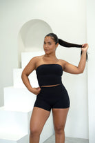 Woman wearing a black tube top and high-waisted shorts standing against a white background.