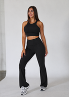 A woman modeling a black athletic two-piece set consisting of a halter crop top and high-waisted flared leggings, paired with silver sneakers, in a studio setting.